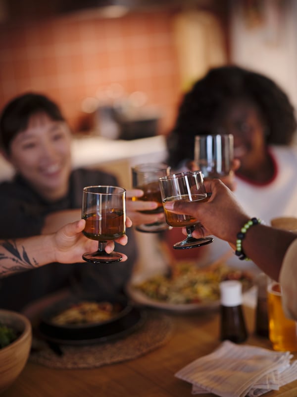 Four people at a table, each holding an IKEA 365+ goblet filled with drinks, reaching towards one another for a toast.