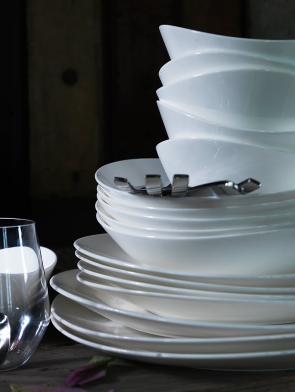 Various sizes of white plates and white serving bowls are stacked on top of each other next to a glass.