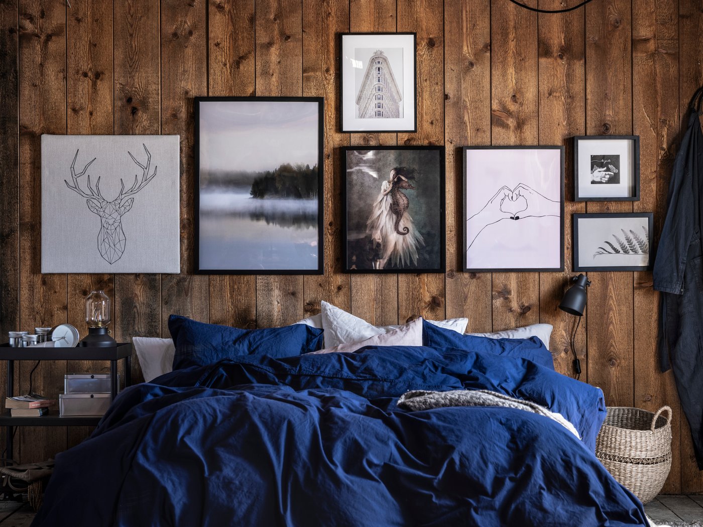 A bed with bed linen in blue and other colours stands in front of a wooden wall decorated with various works of art.