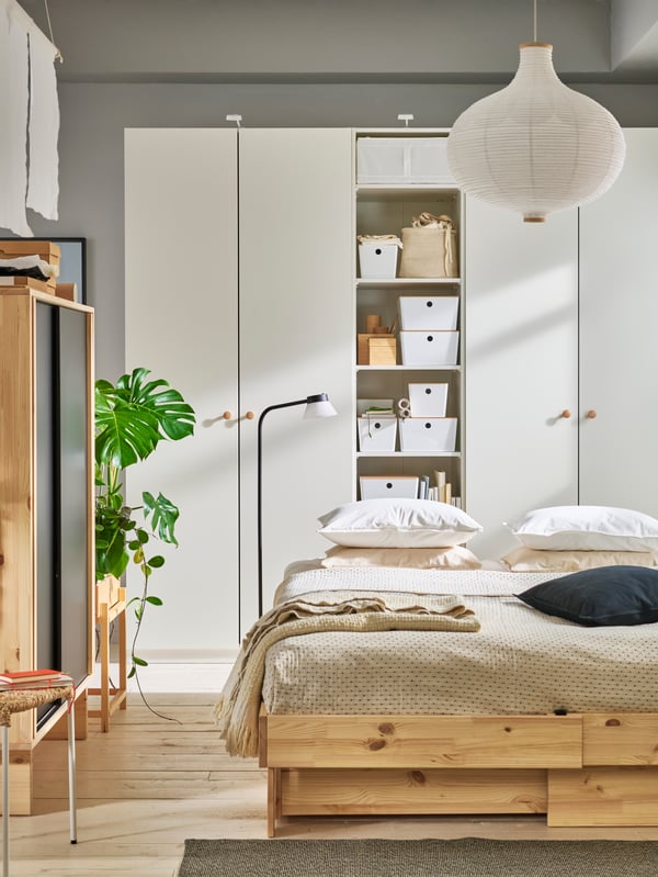 A sunny bedroom with a natural wood bed and beige bedding. A combination of white PLATSA wardrobes are behind the bed.