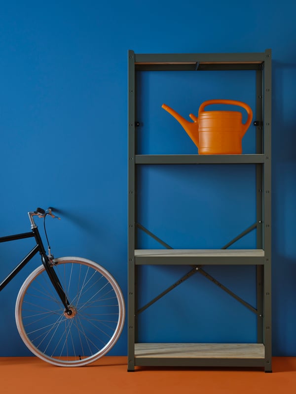 A gray-green BROR shelving unit, holding an orange watering can by a dark blue wall. Next to the shelving unit is a bicycle.