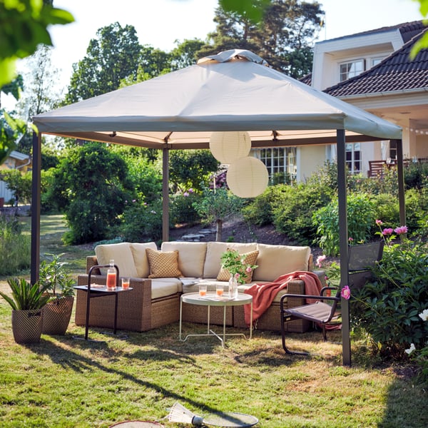 A SOLLERÖN corner outdoor sofa with beige cushions and a black armchair under a HIMMELSÖ gazebo on a sunny garden lawn.