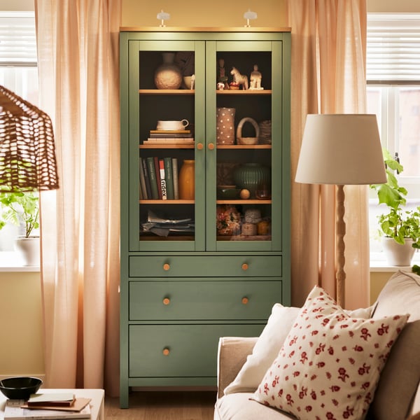 A KINNAHULT floor lamp stands near a HEMNES glass-door cabinet with 3 drawers standing against a wall between two windows.