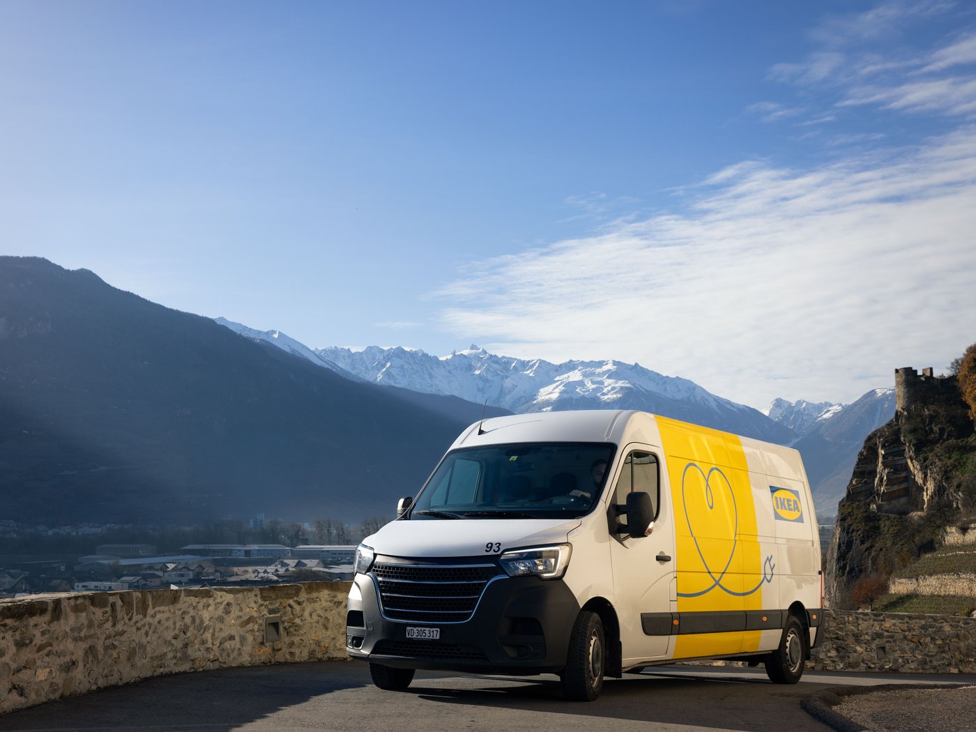 Et eldrevet køretøj med en IKEA levering på en vej i Schweiz. Himlen er blå, og der er en delvist sneklædt bjergkæde i baggrunden.
