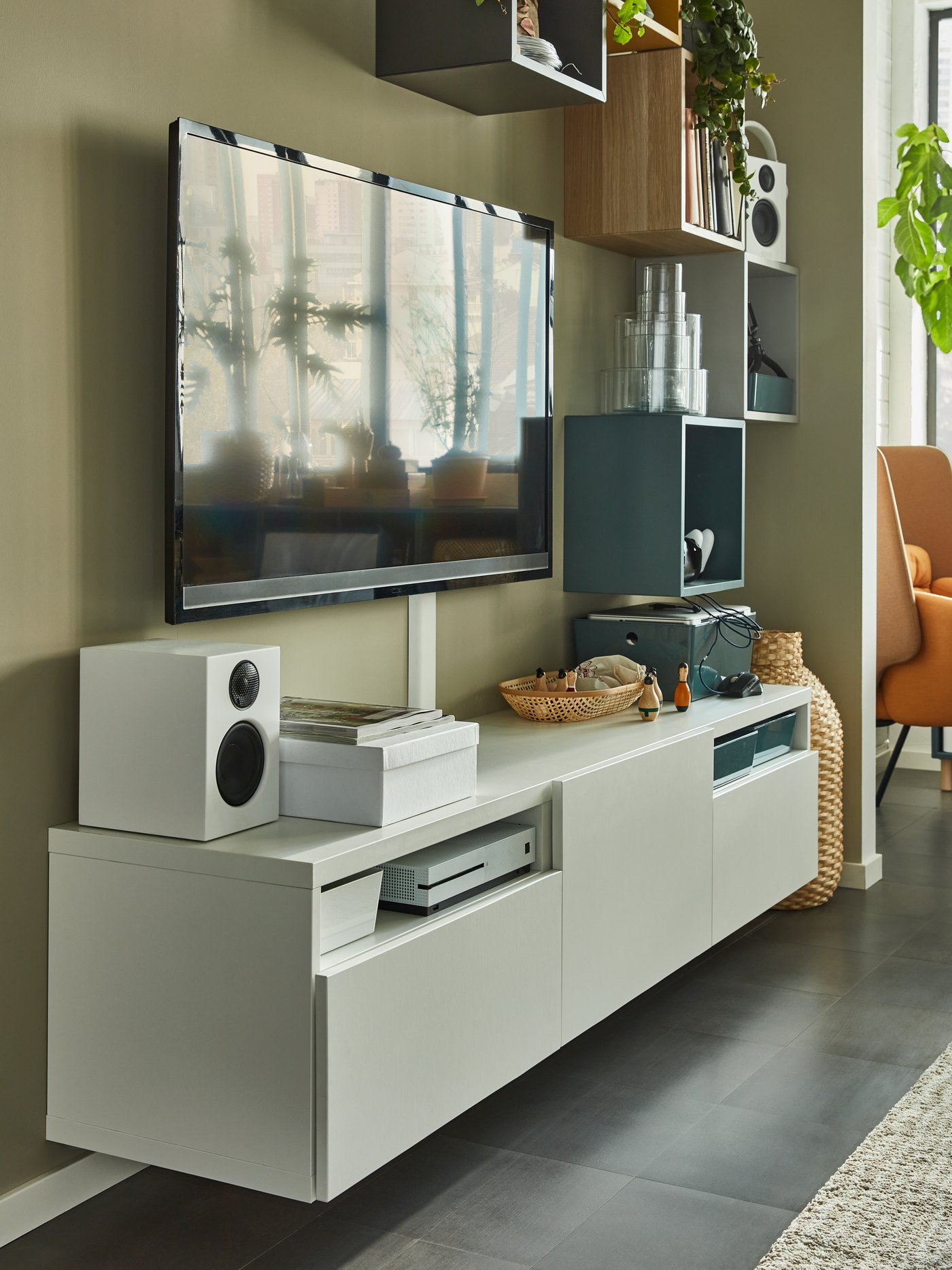 Un salon moderne avec une télévision à écran plat au-dessus d’un meuble TV blanc, entouré de livres, objets déco et une plante près de la fenêtre baignée de lumière naturelle.
