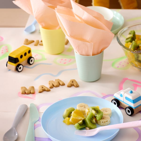 A table with pastel coloured KALAS plates and cutlery, plastic cups with napkins and “mama” spelled out with letter biscuits.