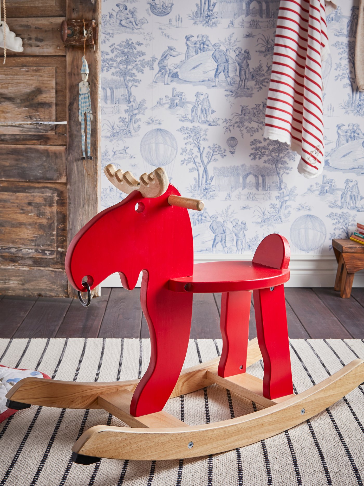 An EKORRE rocking-moose on a striped rug. Clothes hanging on a wall next to an old-looking wooden door that’s open.
