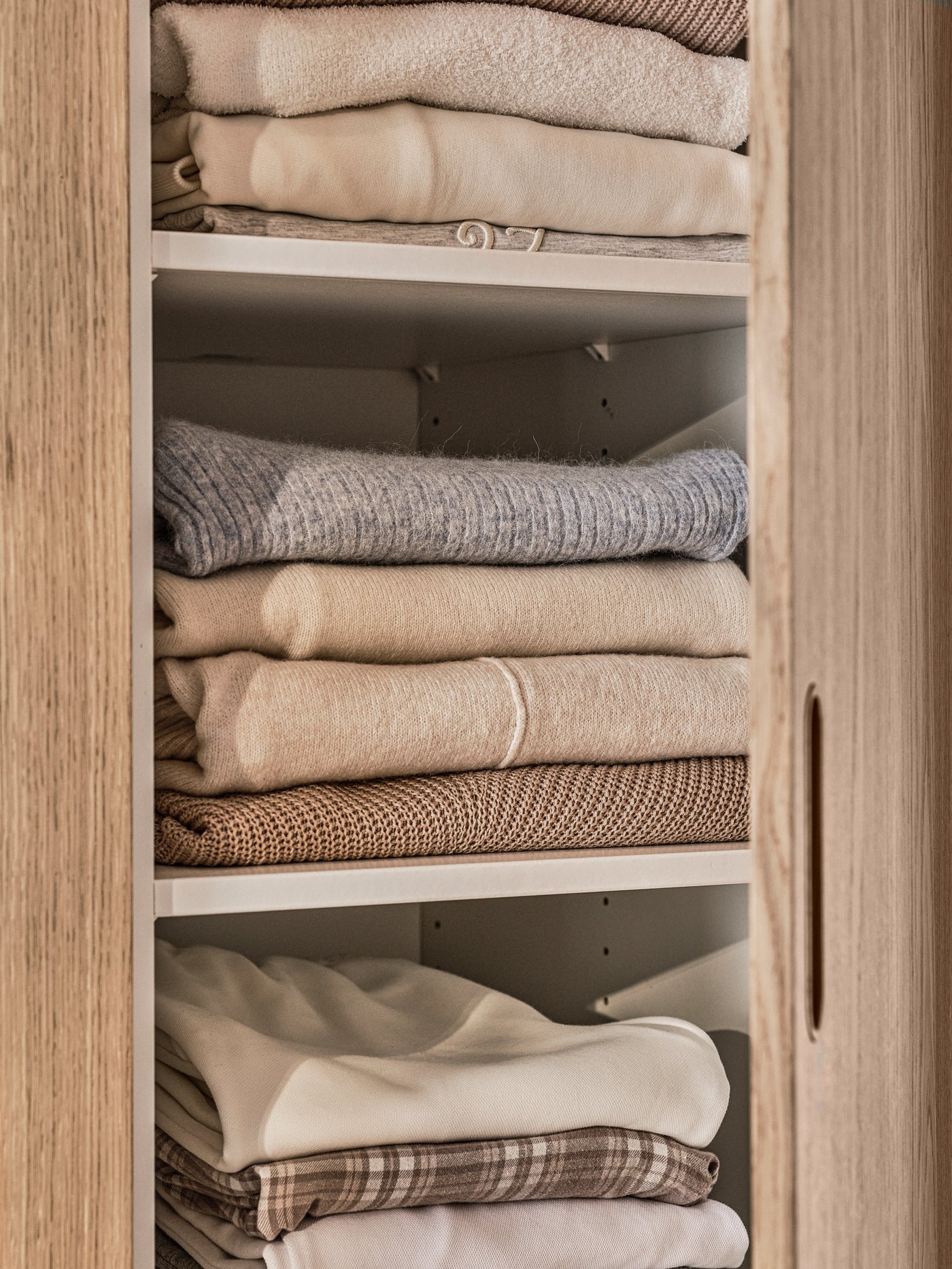 Un clóset PAX/TONSTAD con una puerta abierta que muestra tres estantes, cada uno de los cuales contiene montones de ropa doblada de color claro.