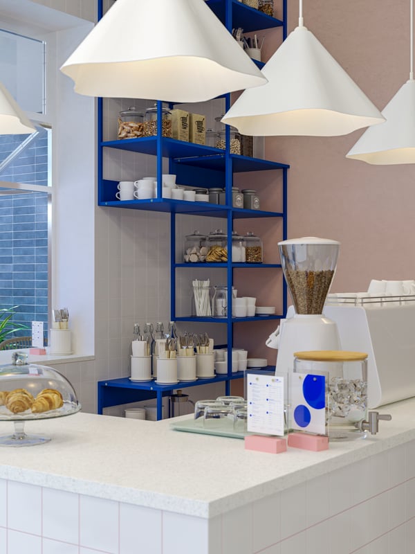 Varias lámparas de techo ZEBRASÄV de plástico de color blanco por encima de la barra de una cafetería con varios utensilios para servir encima y unas estanterías abiertas PLATSA de color azul.