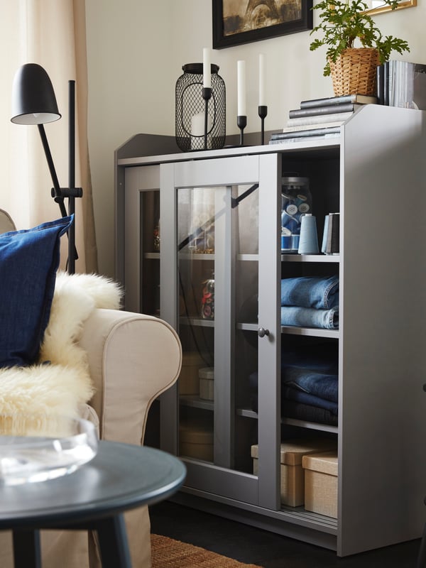 Folded jeans and boxes stored inside a grey glass-door cabinet. A light beige sofa and a black side table are beside.