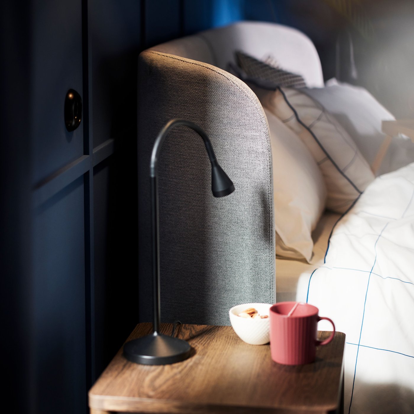 The head of a TUFJORD upholstered bed with a TONSTAD bedside table beside it which has a black NÄVLINGE LED work lamp on top.