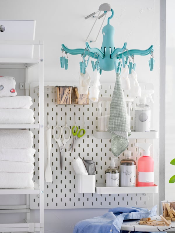 El tablero de clavijas SKÅDIS blanco con ganchos y estantes sostienen las pinzas de ropa, artículos de baño y artículos de lavandería más pequeños.