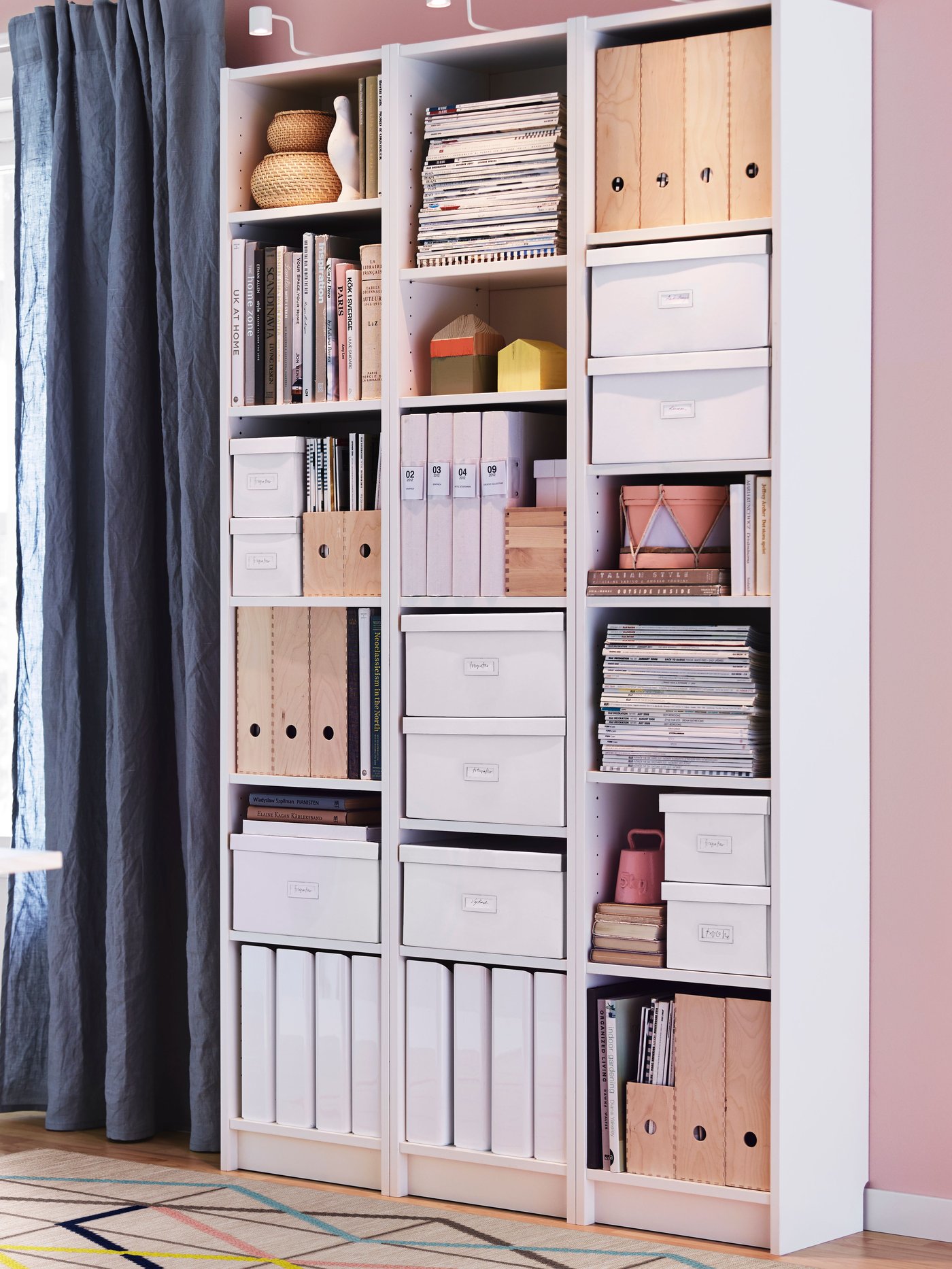 Three white BILLY bookcases with open shelves filled with boxes, organisers and books against a pink wall in a bright room.