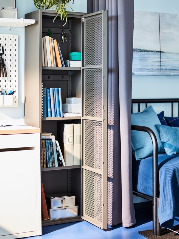 A grey IVAR cabinet with an open mesh door between a white desk and a black day-bed. Books on the shelves.