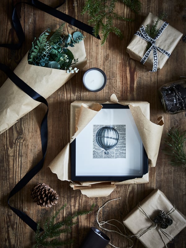 A black SANNAHED picture frame with wrapping paper around it next to an in/outdoor green SMYCKA artificial bouquet on the table.