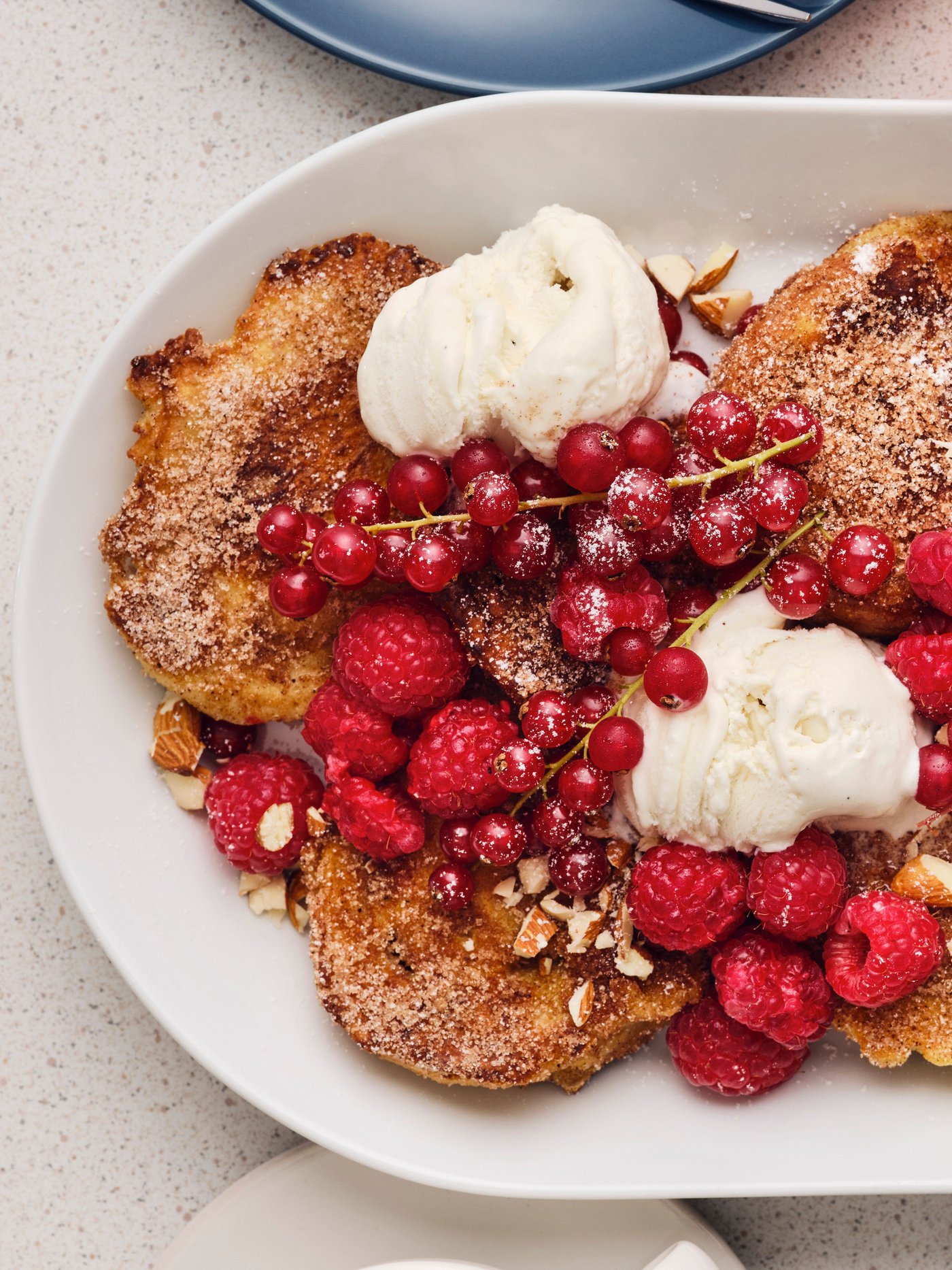 Un plat ovale blanc contient plusieurs morceaux de pain perdu avec de la crème glacée, des framboises fraîches, des groseilles et des amandes.