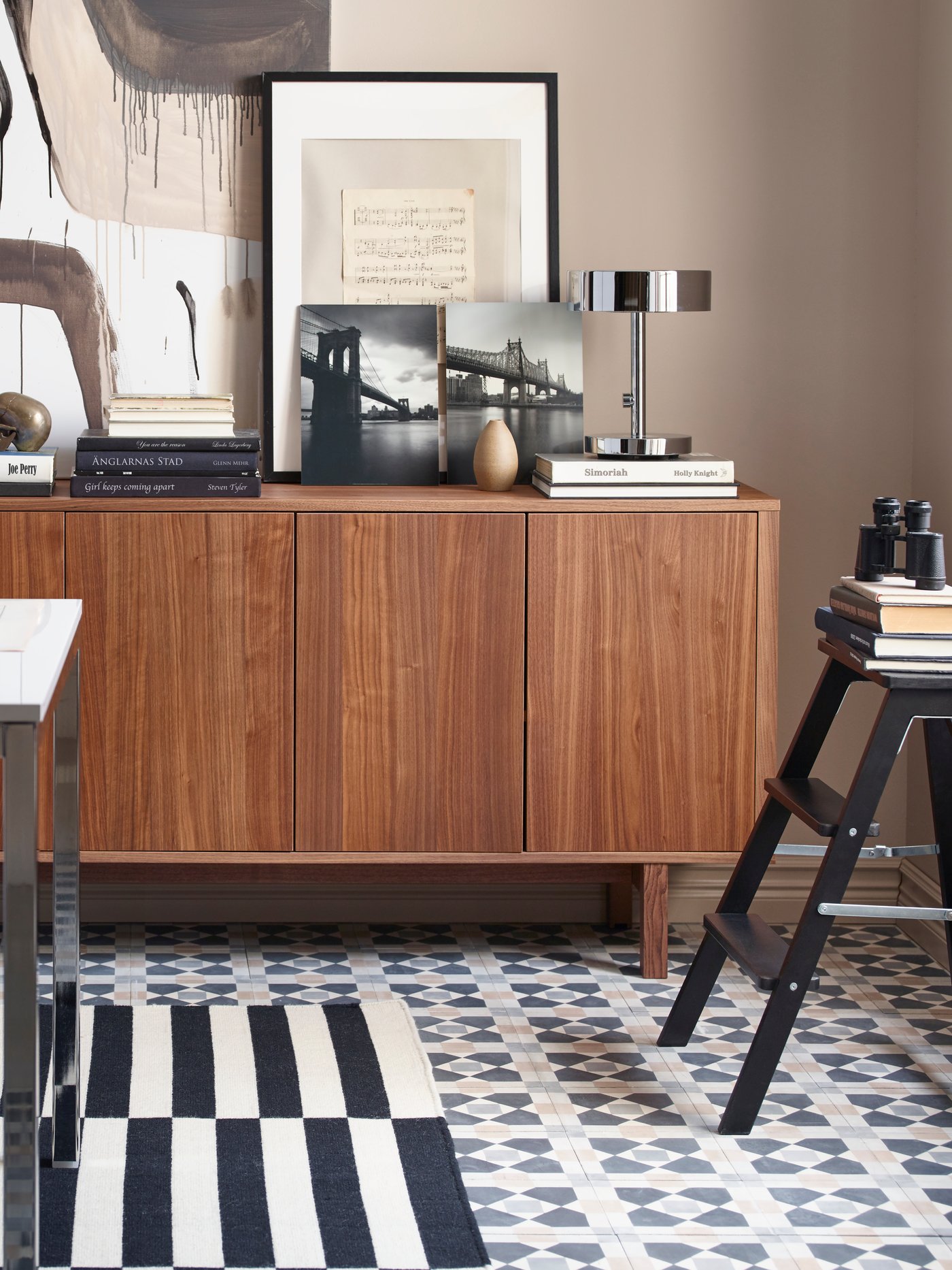 Un intérieur moderne avec un buffet en bois contre un mur beige, décoré de cadres, livres, lampe et vases, à côté d’un escabeau noir portant des livres, sur un tapis rayé noir et blanc.