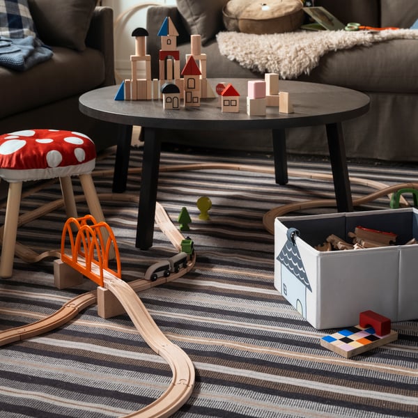A STOCKHOLM 2025 coffee table, toys, and a FLISAT children's stool with a cover on it are placed on a RÅVAROR rug in a living room.