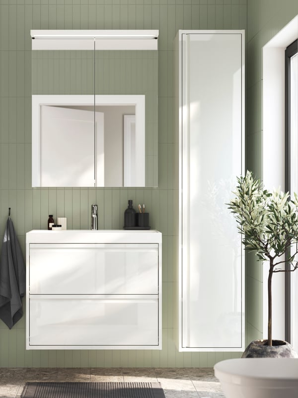 Meuble lavabo ÄNGSJÖN blanc dans une salle de bains carrelée verte, à côté d’une armoire haute et d’un miroir avec éclairage intégré.