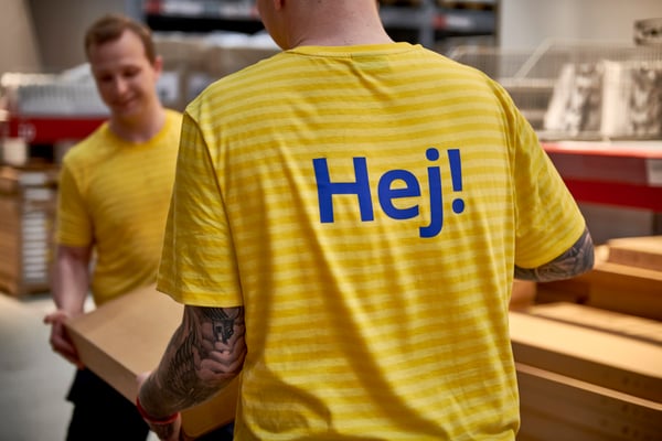 Deux employés d’IKEA dans un entrepôt, reconnaissables à leurs vêtements de travail rayés jaunes avec « Hej ! » dans le dos ; Un employé porte une planche de bois, avec des étagères remplies de produits en arrière-plan.