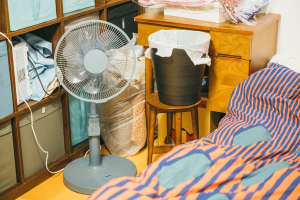 寝具と収納棚の間に、扇風機とゴミ箱が置かれている。