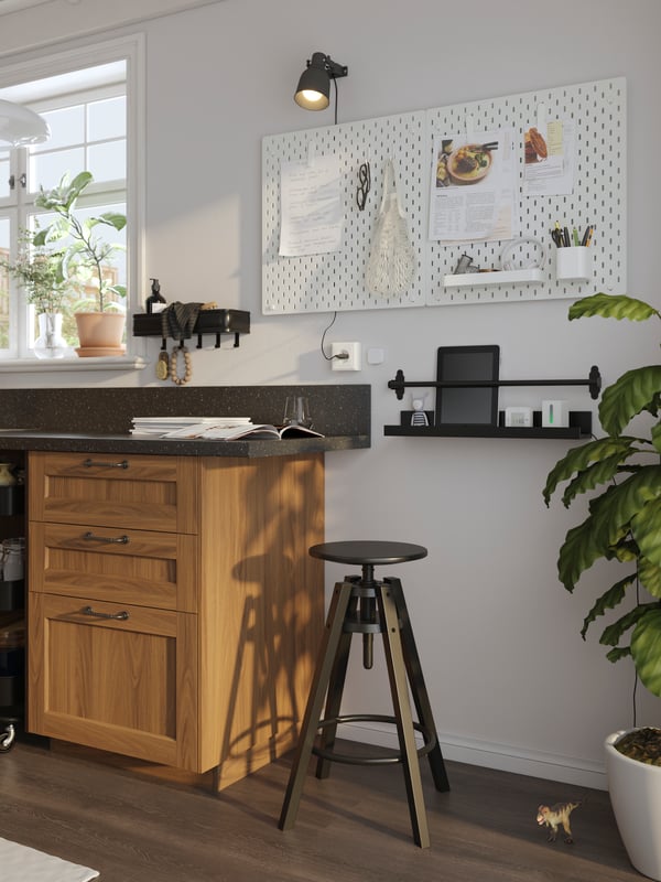 A black DALFRED bar stool at the end of a kitchen counter with a black shelf and white memo board on the wall behind it.