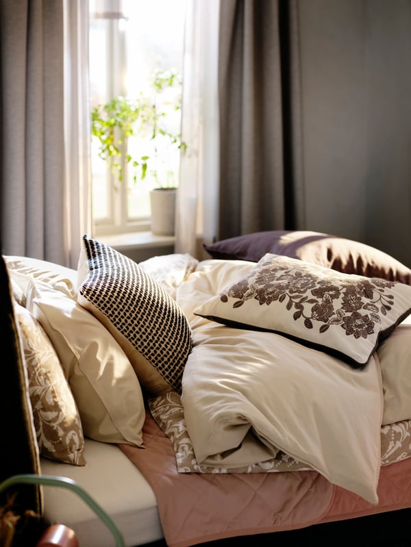 Una cama con ropa de cama ÄNGSLILJA y cojines encima de plumón. Detrás de la cama hay una ventana con una cortina VILBORG.