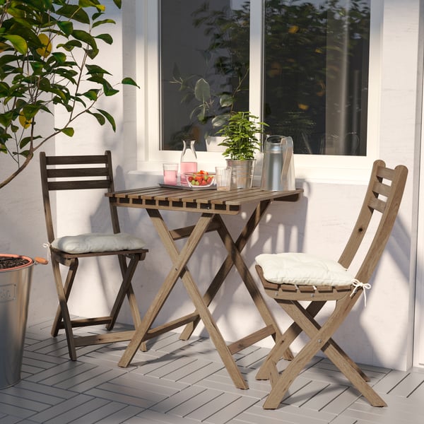 A wooden outdoor table holding a carafe, drinking glass, a bowl of fruit and a plant, two outdoor chairs with cushions.