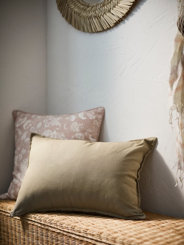 A rectangular cushion in a beige GURLI cover in front of a square cushion in a light pink IDALINNEA cover on a rattan bench.