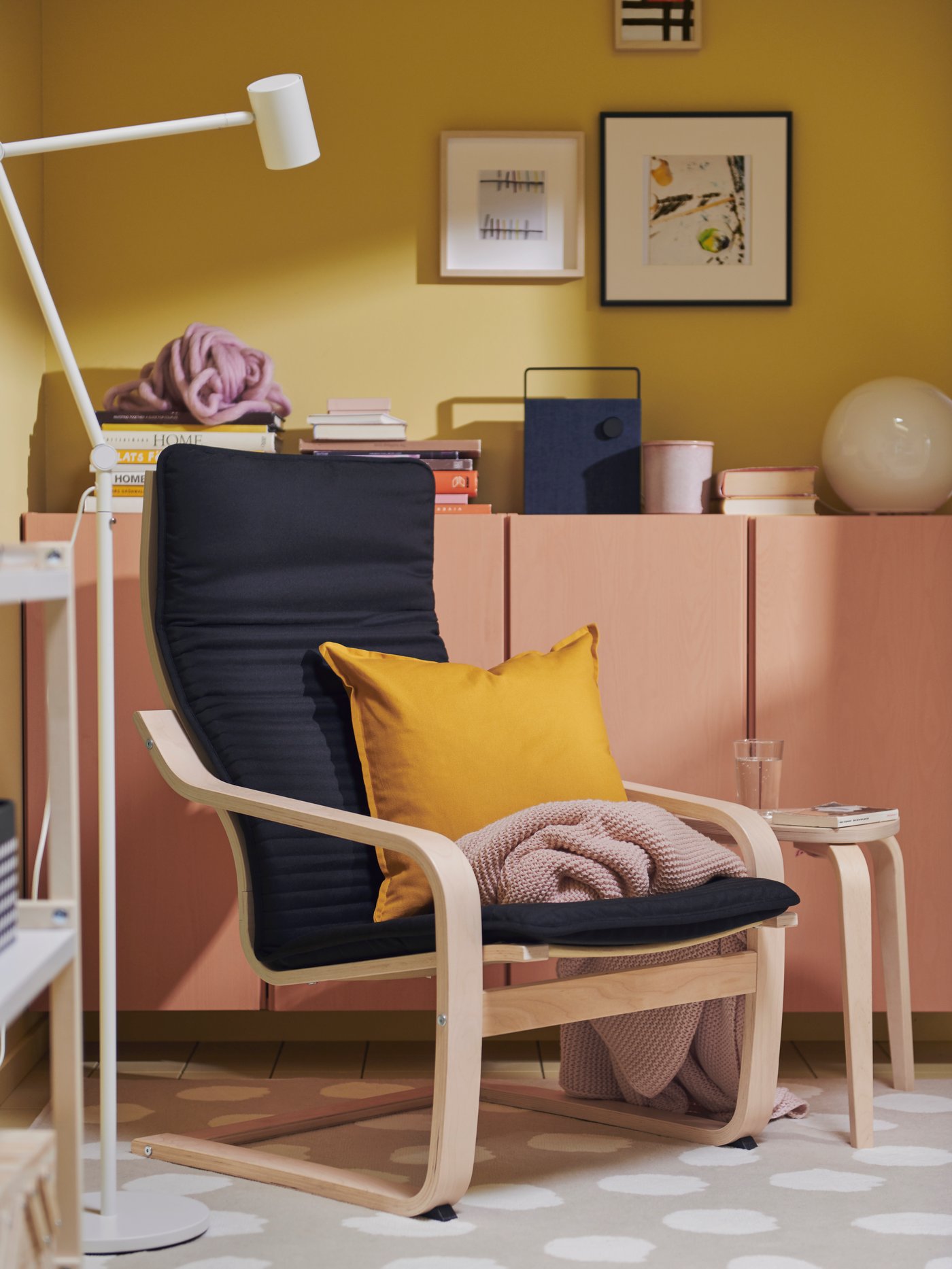A white NYMÅNE floor/reading lamp and a black POÄNG armchair in birch veneer next to IVAR cabinets and a Bluetooth speaker.
