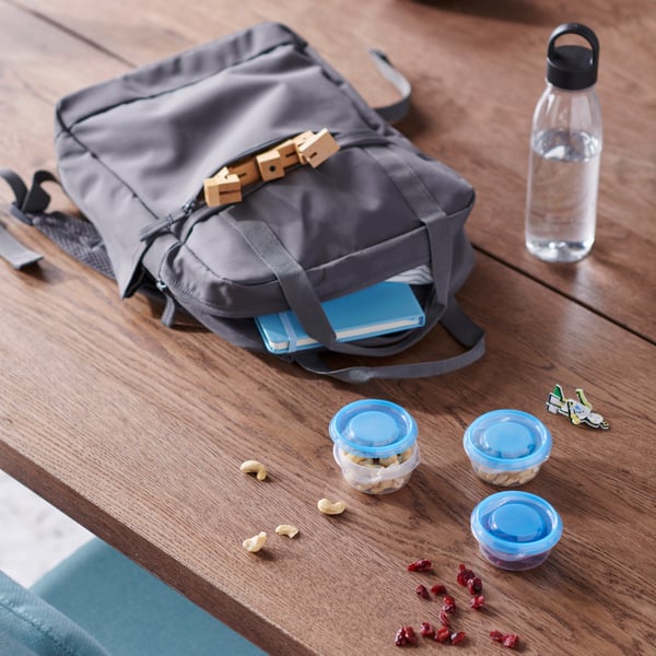 Sac STARTTID gris avec de petits contenants pour collations bleus dont le contenu se renverse sur une table en bois. Une bouteille d’eau transparente est juste à côté.