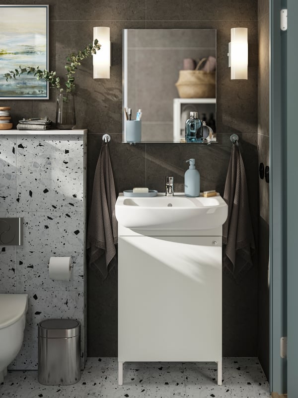 A NYSJÖN wash-basin cabinet stands under a white wash-basin with light grey-blue accessories on top and a mirror on the wall.