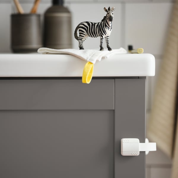Hračka zebra stojí na žínce umístěné na šedé skříňce ENHET v koupelně s bílými obklady.