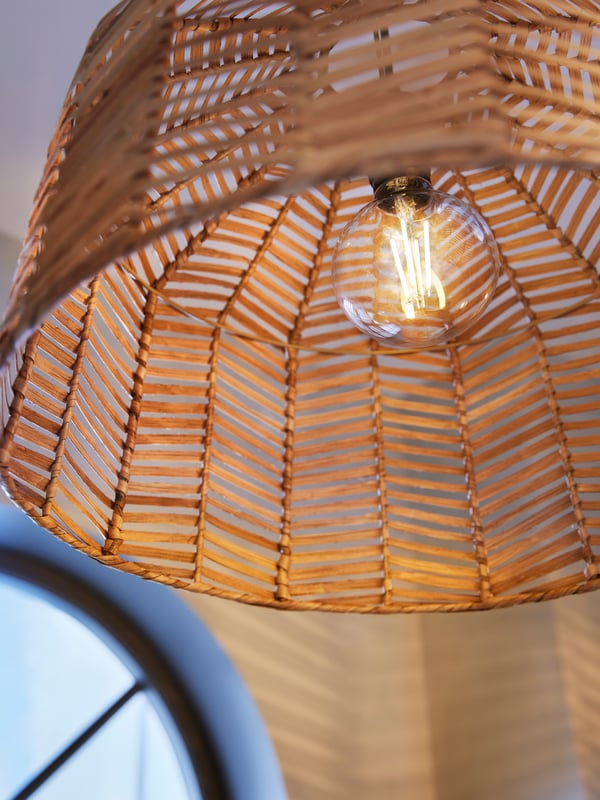 A lit TRÅDFRI E27 470 lumen LED bulb inside a rattan KAPPELAND pendant lamp shade. The light casts a pattern on the wall.