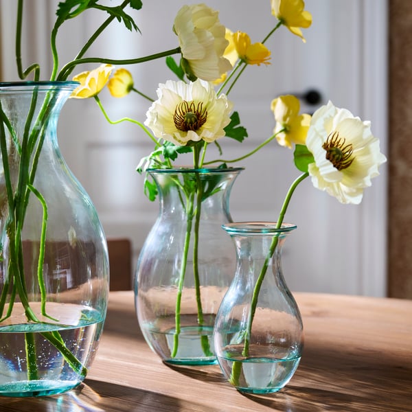 Three BERGKÖRSBÄR vases in clear glass, placed on a wooden table, each holding water and yellow flowers with green stems.