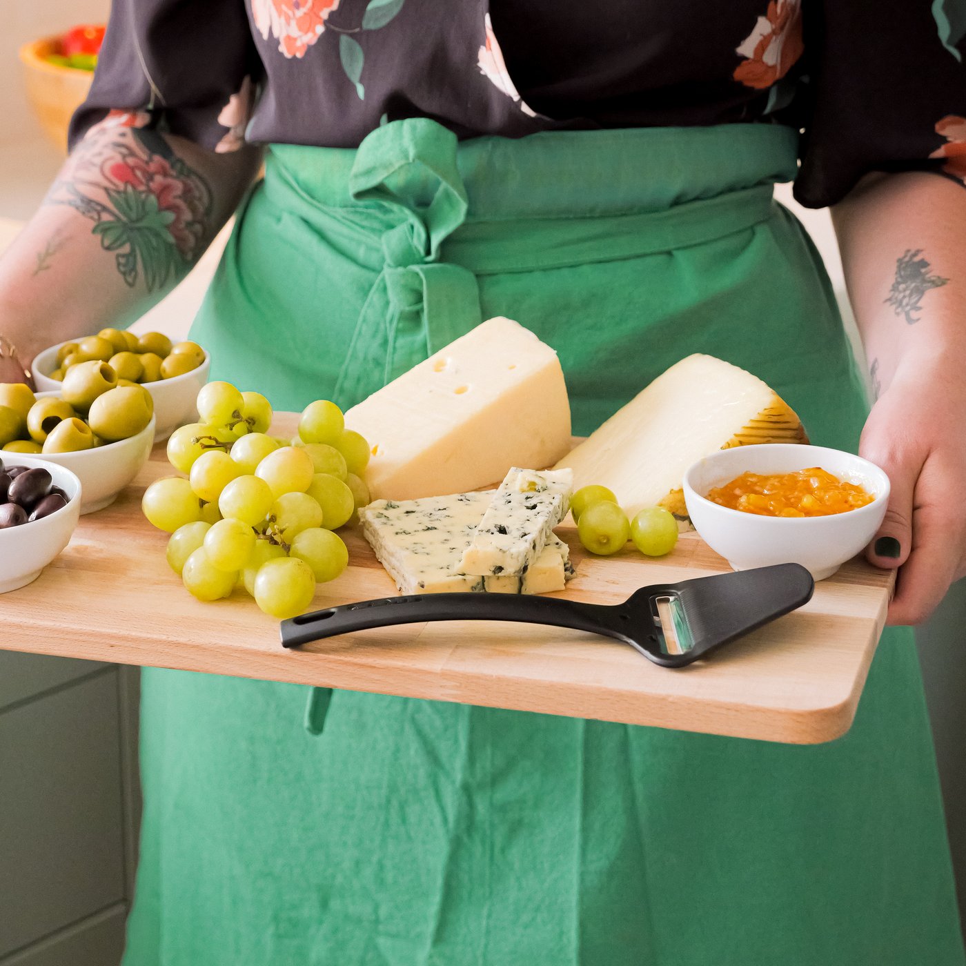 A woman in an apron carries a PROPPMÄTT chopping board, filled with cheese, fruits, and olives in small IKEA 365+ bowls.