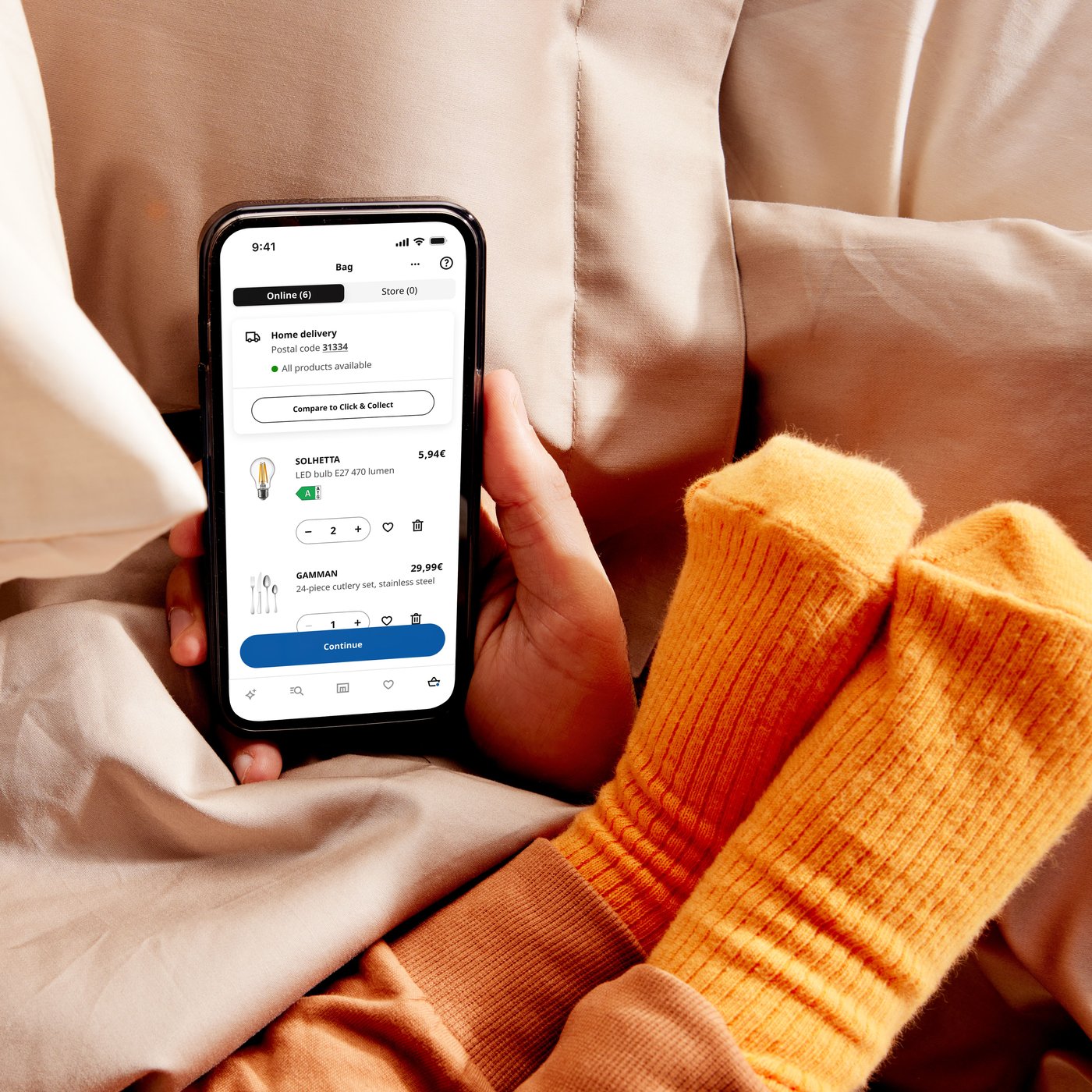 On a bed, a hand is holding a smartphone displaying the IKEA app, while a small child's feet with orange socks rests on the persons arm.