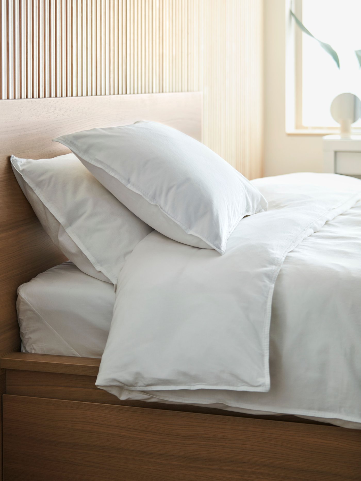 Close-up of a bed with sheets and two fluffy pillows in crisp white, the daylight shining through a window in the back.