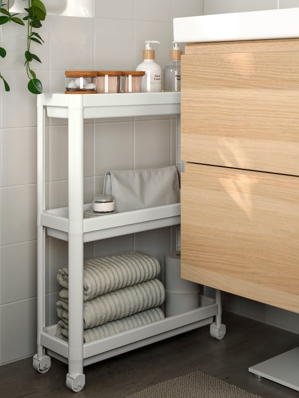 VESKEN trolley with dispensers and bottles, wedged between a white-tiled wall and wash-stand with wooden drawer fronts.