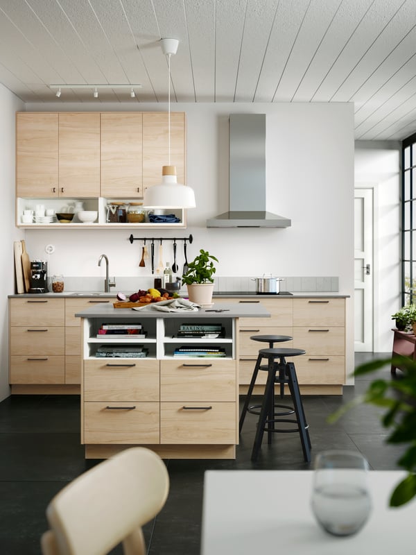 Un îlot de cuisine comporte des tiroirs aux façades FRÖJERED en bambou clair, une grande suspension blanche au-dessus et deux tabourets noirs.