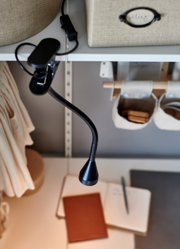 Faretto a LED con morsetto NÄVLINGE nero fissato a una mensola al di sopra di una scrivania bianca su cui ci sono alcuni quaderni e una penna - IKEA.