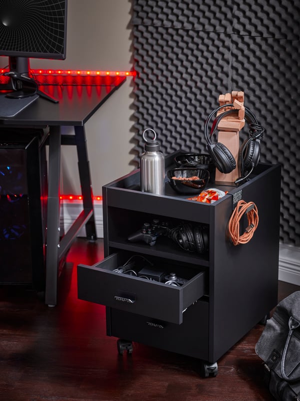 An IKEA UPPSPEL black short drawer unit on castors next to a black gaming desk lit up with red light strips, with a steel water bottle, black headphones and other gaming accessories on top.
