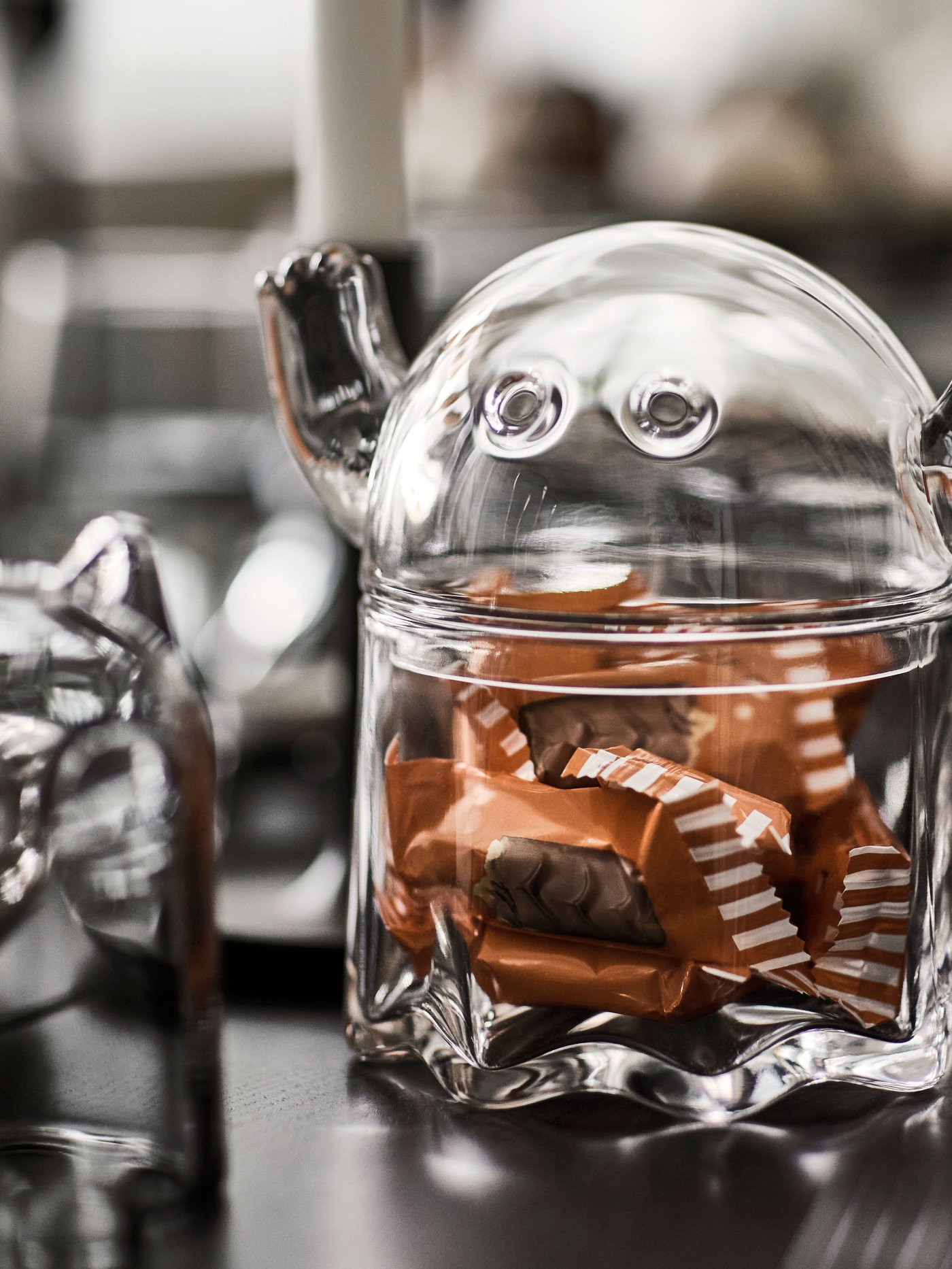A KUSTFYR glass container shaped like a ghost, containing sweets wrapped in brown wrappers, on a polished table.