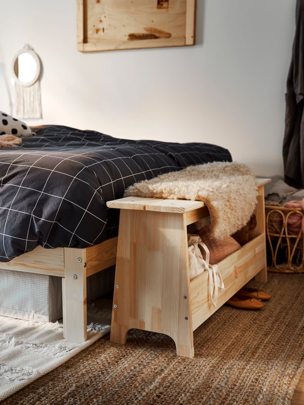 Un banc en pin avec rangement PERJOHAN, un tapis posé dessus et divers objets rangés à l’intérieur, sur un tapis de jute au pied d’un lit.