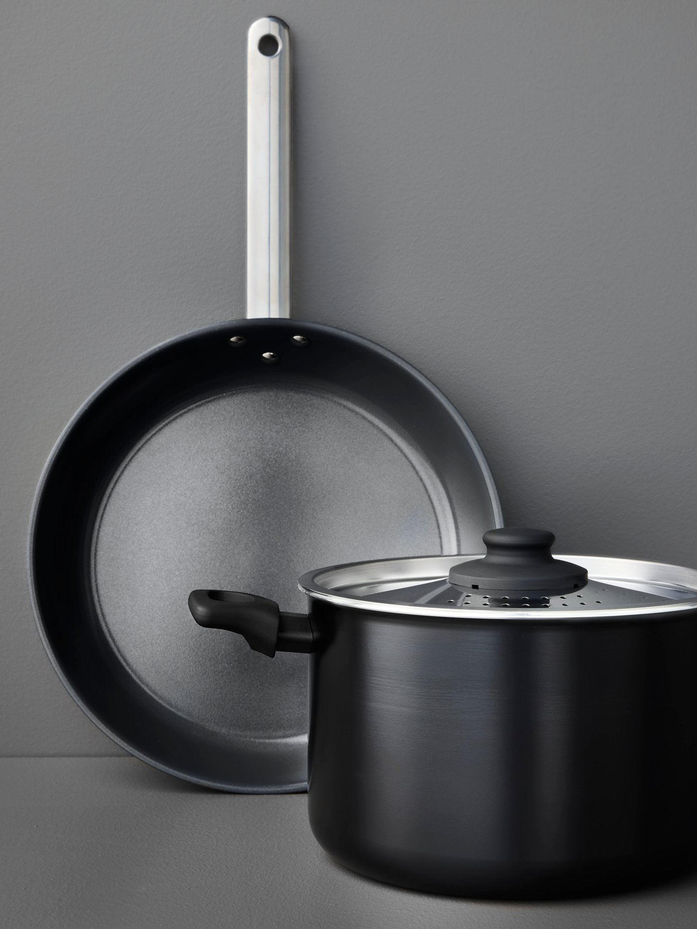 A black HEMLAGAD frying pan and a HEMLAGAD pot with lid, both with non-stick coating, are placed on a grey surface.