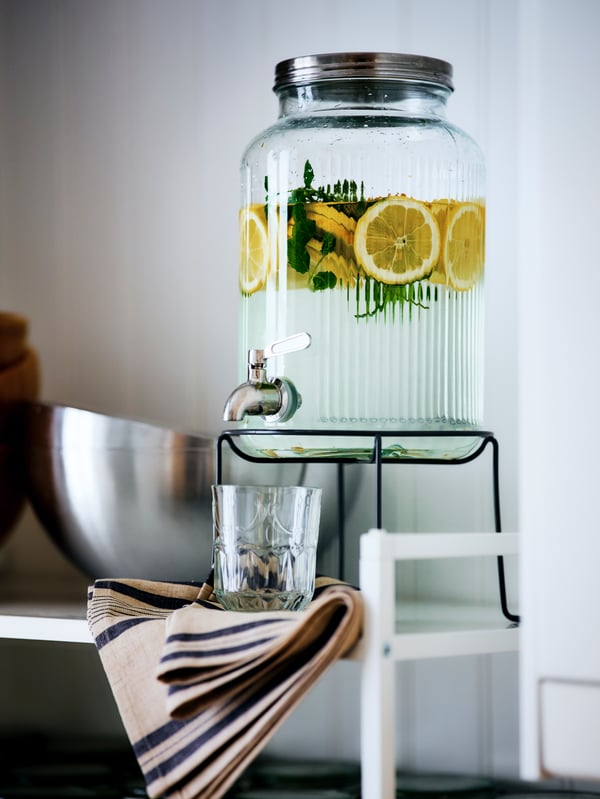 A VARDAGEN jar with tap filled with lemon slices and herbs held in a stand on a white shelf, with a glass under the tap.