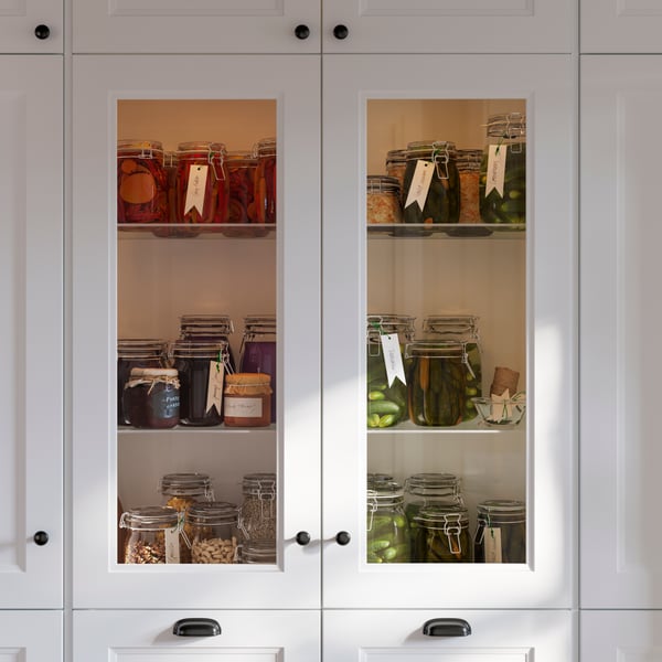 Une armoire de cuisine blanche à porte vitrée montre des bocaux de sauce, de fruits et de légumes en conserve, tous bien organisés.