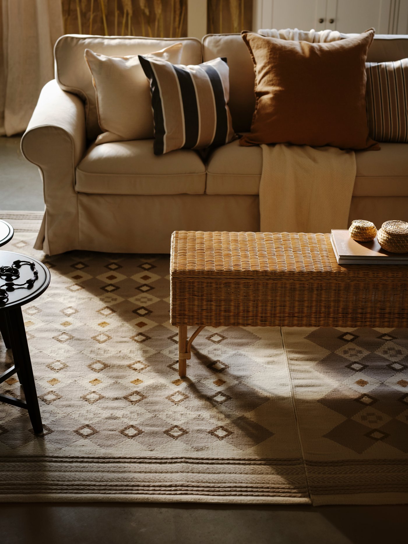 Ein Sofa mit verschiedenen Kissen in erdigen Tönen und ein cremefarbener HUMLEMOTT Überwurf liegen auf einem beigen EKTORP Sofa. Unter dem Sofa liegt ein VÄGNÄT Teppich in grau und elfenbeinweiß.