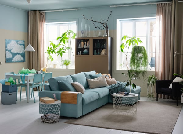 Ein Wohnzimmer mit einem grossen Sofa, das auf einem Teppich in der Mitte des Raumes steht, dahinter eine Essplatz-Gruppe. Zwei grosse Fenster vor denen Pflanzen stehen mit langen Vorhängen lassen viel Licht in den Raum. 