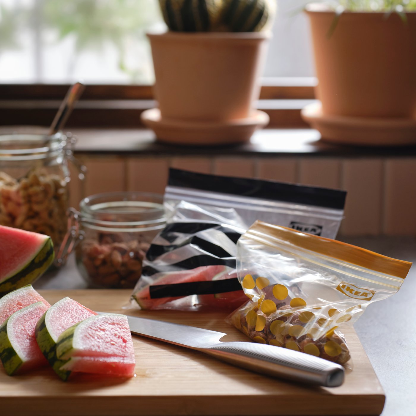 Slices of watermelon, a knife and two different sized ISTAD plastic resealable bags are on a cutting board in a kitchen.
